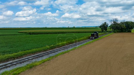 Eine einzige Lokomotive durchquert Eisenbahngleise, die an ein gepflügtes Feld und grüne Feldfrüchte unter einem weiten Himmel mit aufgedunsenen Wolken grenzen.
