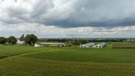 Une ferme agricole paisible se trouve sous un ciel dramatique, où les nuages denses laissent entrevoir le temps changeant sur le paysage luxuriant.