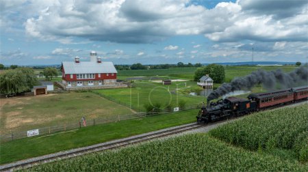 Photo for Ronks, Pennsylvania, August 15, 2023 - A vintage locomotive steams by a picturesque farm with its red barn and pond, adding a touch of historical charm to the rural landscape. - Royalty Free Image