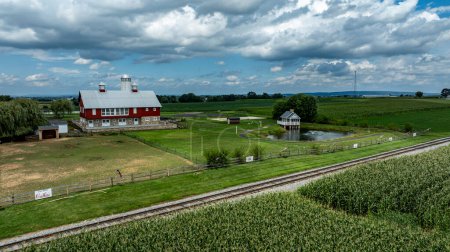 Photo for Ronks, Pennsylvania, August 15, 2023 - Dominating this pastoral scene is a classic red barn with a serene pond and gazebo, set against the rich tapestry of flourishing cornfields. - Royalty Free Image