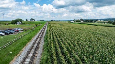 Parallel to a train track, this image shows a flourishing cornfield beside a parking area, capturing a slice of rural Americana under a dynamic sky.