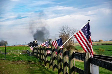 Historyczny pociąg parowy wznosi się wzdłuż wsi, otoczony amerykańskimi flagami na drewnianym płocie, przywołując uczucie nostalgii w Ameryce i historycznej podróży.