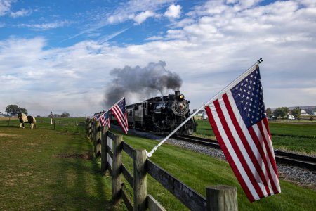 A vintage steam train chugs along the countryside, flanked by American flags on a wooden fence, evoking a sense of nostalgic Americana and historic travel.