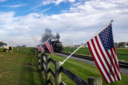 Ein historischer Dampfzug tuckert durch die Landschaft, flankiert von amerikanischen Flaggen an einem Holzzaun, der ein Gefühl nostalgischer Americana und historischer Reisen heraufbeschwört..