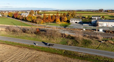 Un buggy Amish serein parcourt une route de Lancaster, en Pennsylvanie, flanqué des couleurs vibrantes de l'automne et de vastes terres agricoles, témoignage du riche patrimoine agricole des régions..
