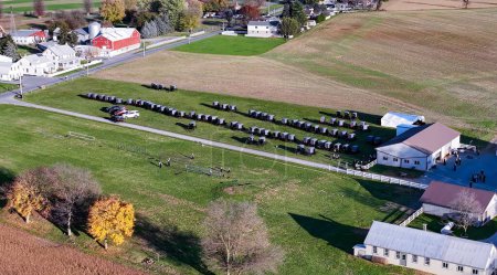 Eine Luftaufnahme einer ländlichen Gemeinschaftsveranstaltung, bei der reihenweise Pferdewagen neben einem Bauernhof geparkt sind, die den Geist der Zweisamkeit und Tradition verkörpern. Amische Hochzeit