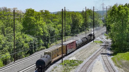 An engaging aerial view of a freight train carrying diverse cargo winding through rural tracks surrounded by lush greenery, under a bright sunny sky.