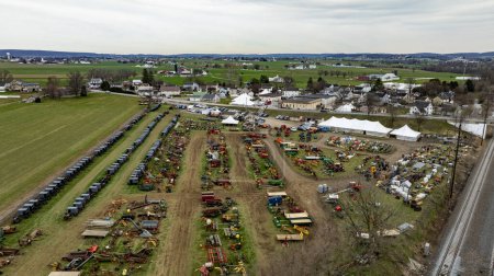 This Aerial image of an Amish Mud Sale, shows a comprehensive display of colorful farm machinery and equipment spread across a rural exhibition field with tents and visitors.