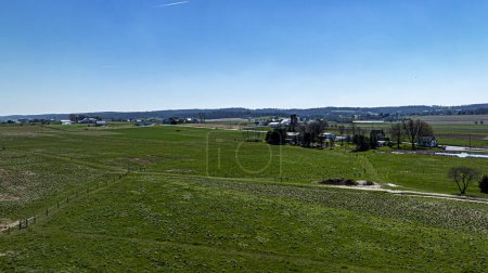 An expansive view from above showcasing lush green pastures segmented by fencing, with a detailed farmstead and distant hills under a brilliant blue sky.