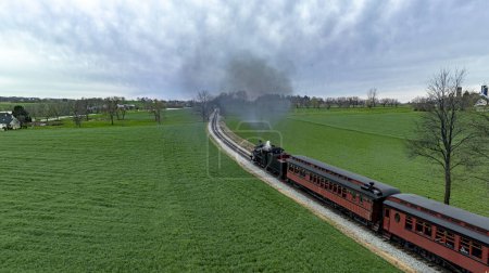 Captivating aerial photo of a historic steam train emitting a dense smoke plume as it winds through vibrant green fields, under a dramatic overcast sky.