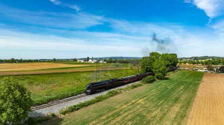 Photo for A striking aerial view showcasing a historic train cutting across scenic farmlands, with mixed agriculture fields and a backdrop of blue sky. - Royalty Free Image