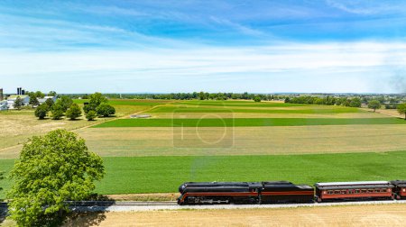 Photo for Stunning aerial view of a classic train passing through expansive fields, weaving its path between lush greenery and farmland under a clear sky. - Royalty Free Image