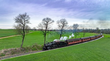 Photo for Eye-catching aerial image of a classic steam locomotive pulling red passenger cars through a lush green countryside, with trees and a cloudy sky adding to the scenic beauty. - Royalty Free Image