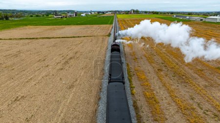 Photo for Dramatic top-down view of a steam train emitting a thick cloud of smoke as it travels alongside freshly harvested fields, highlighting the contrast between technology and nature. - Royalty Free Image