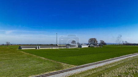 Fesselnde Luftaufnahme eines ruhigen Bauernhofes mit modernen landwirtschaftlichen Gebäuden und lebendigen grünen Feldern neben einer Eisenbahnstrecke, unter einem klaren blauen Himmel.