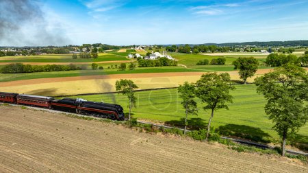 An evocative aerial view of a steam train puffing through picturesque rural farmlands, marked by vividly colored fields and pastoral settings.