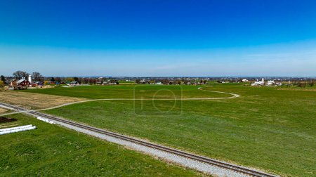 Esta fotografía aérea muestra un paisaje rural amplio e idílico con exuberantes campos verdes, una carretera curva y una vía férrea, sobre un telón de fondo de un pequeño pueblo y un cielo azul claro..
