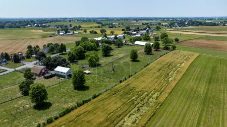 La fotografía aérea muestra un paisaje mixto de áreas residenciales y agrícolas, con casas, edificios agrícolas y campos de varios colores bajo un cielo azul claro..