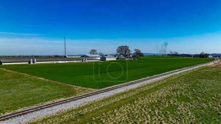 Captivating aerial view of a serene farm with modern agricultural buildings and vibrant green fields next to a railroad track, under a clear blue sky.