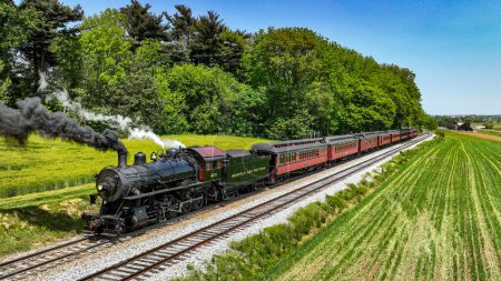 Photo for A classic Norfolk and Western steam locomotive hauls vintage passenger cars through a picturesque rural setting. The train, belching thick black smoke, travels alongside vibrant green fields - Royalty Free Image