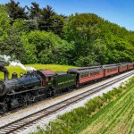 A classic Norfolk and Western steam locomotive hauls vintage passenger cars through a picturesque rural setting. The train, belching thick black smoke, travels alongside vibrant green fields