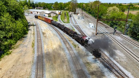 An aerial powerful vintage steam train moves through an industrial rail yard, emitting steam as it passes warehouses and alongside other rail cars under a clear sky.