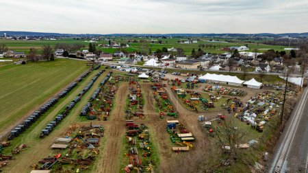 This Aerial image of an Amish Mud Sale, shows a comprehensive display of colorful farm machinery and equipment spread across a rural exhibition field with tents and visitors.