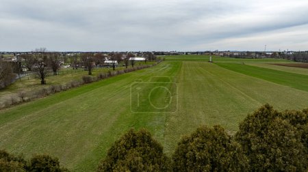 This drone-captured image showcases a vast rural landscape with distinct green fields, a few clusters of trees, and a small town in the background under a cloudy sky.