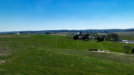 Una vista amplia desde arriba que muestra exuberantes pastos verdes segmentados por esgrima, con una granja detallada y colinas distantes bajo un cielo azul brillante.