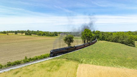 Photo for Captivating aerial view of a vintage steam train traveling through a vast rural landscape, with plowed and green fields under a blue sky. - Royalty Free Image