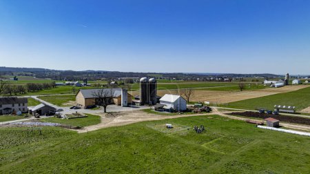 Foto aérea de un concurrido complejo agrícola, con múltiples graneros y silos rodeados de verdes pastos, equipo agrícola en uso, y un grupo de personas reunidas, bajo un cielo despejado.