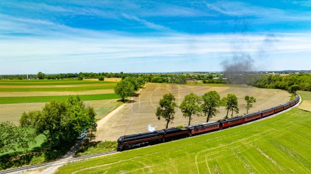 Photo for Aerial image of a classic steam train curving through the countryside, emitting smoke above vibrant and plowed fields under a blue sky. - Royalty Free Image
