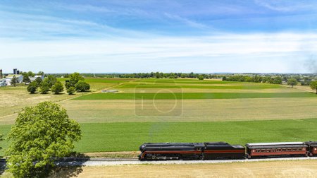 Superbe vue aérienne d'un train classique traversant de vastes champs, tissant son chemin entre verdure luxuriante et terres agricoles sous un ciel dégagé.