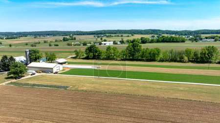 Eine ausgedehnte Luftaufnahme, die die heitere Schönheit eines ländlichen Bauernhofes einfängt und eine Mischung aus gepflügten Feldern, grünen Feldfrüchten und landwirtschaftlichen Gebäuden unter einem klaren blauen Himmel zeigt.