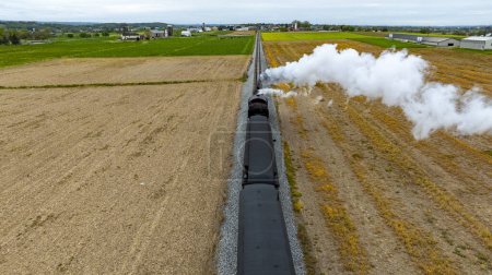 Dramatischer Blick von oben auf einen Dampfzug, der eine dicke Rauchwolke ausstößt, während er an frisch abgeernteten Feldern vorbeifährt, was den Kontrast zwischen Technologie und Natur unterstreicht.