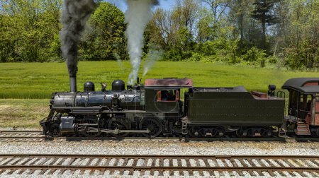 Photo for A detailed close-up image of a vintage steam locomotive, Number 89, showing the engine emitting thick smoke and steam as it operates on the tracks, surrounded by lush greenery on a bright sunny day. - Royalty Free Image