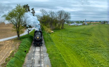 Photo for Atmospheric shot of a vintage steam train chugging through a rural landscape, emitting thick smoke, with a backdrop of freshly plowed fields and lush greenery. - Royalty Free Image