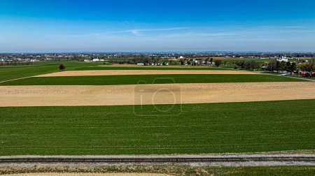 Eine atemberaubende Luftaufnahme, die den lebendigen Flickenteppich landwirtschaftlicher Felder entlang eines kleinen Dorfes einfängt, mit deutlichen grünen und braunen Flecken unter einem klaren blauen Himmel.