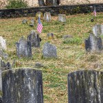 Poignant image of an old cemetery with weathered tombstones and American flags, nestled among fallen autumn leaves, showcasing a solemn tribute to the past.