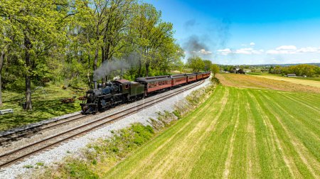 Photo for An enchanting aerial view of a historic steam train emitting a cloud of smoke as it travels through a lush countryside, flanked by vibrant green fields and dense trees under a clear blue sky. - Royalty Free Image