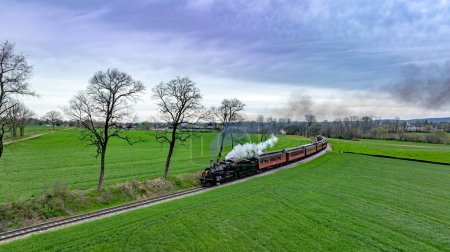 Dramatic aerial capture of a vintage steam train chugging along a curvy track through lush green fields, with a backdrop of dramatic cloudy skies and a distant village.