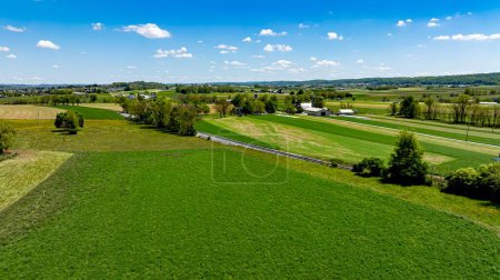Superbe vue aérienne capturant un paysage agricole diversifié avec des champs de différentes nuances de vert, une petite route et des bâtiments lointains sous un ciel parsemé de nuages.