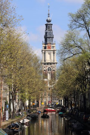 Foto de Amsterdam, Países Bajos - 16 de abril de 2022: Canal en Amsterdam con barcos casa y personas en barcos y la torre de la iglesia de Westerkerk en Amsterdam, Países Bajos el 16 de abril de 2022 - Imagen libre de derechos