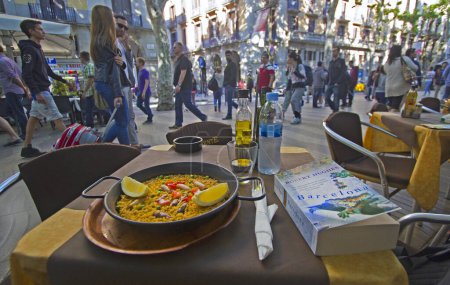 Photo for Barcelona, Spain - May 21, 2015: Plate of Paella and book about Barcelona by Robert Hughes on a table of a restaurant on the Ramblas street in downtown Barcelona, Spain on May 21, 2015. - Royalty Free Image