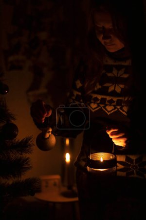 Photo for Ukraine, winter 2022. A woman decorates a Christmas tree in the dark with a candle. - Royalty Free Image