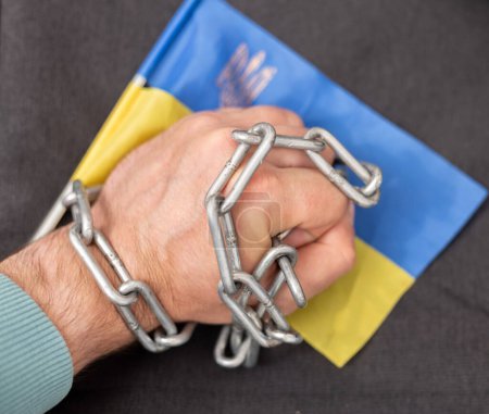 La mano de un hombre enredada en una cadena y la bandera de Ucrania. Prohibición a los ciudadanos de abandonar el país