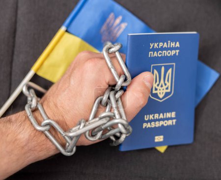 The hand of a man with a Ukrainian passport entangled in a chain and the flag of Ukraine. Ban on citizens leaving the country