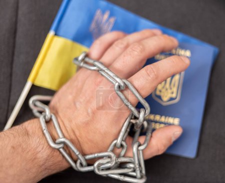 The hand of a man with a Ukrainian passport entangled in a chain and the flag of Ukraine. Ban on citizens leaving the country