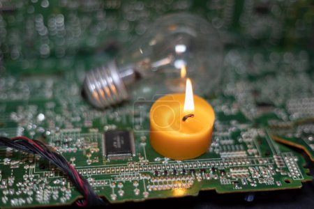 A burning candle against the background of an incandescent lamp and electronic circuit boards. Blackout due to the war in Ukraine