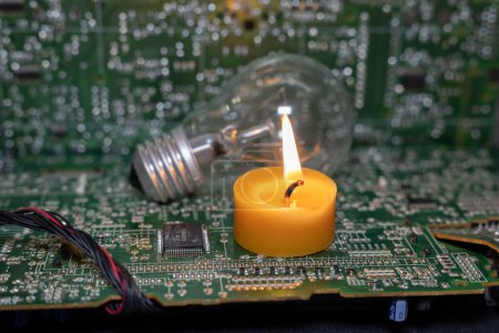 A burning candle against the background of an incandescent lamp and electronic circuit boards. Blackout due to the war in Ukraine
